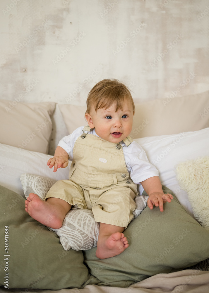 Caucasian baby boy in beige jumpsuit sits on pillows on the bed and looks at the camera Caucasian baby boy in beige jumpsuit sits on pillows on the bed and looks at the camera