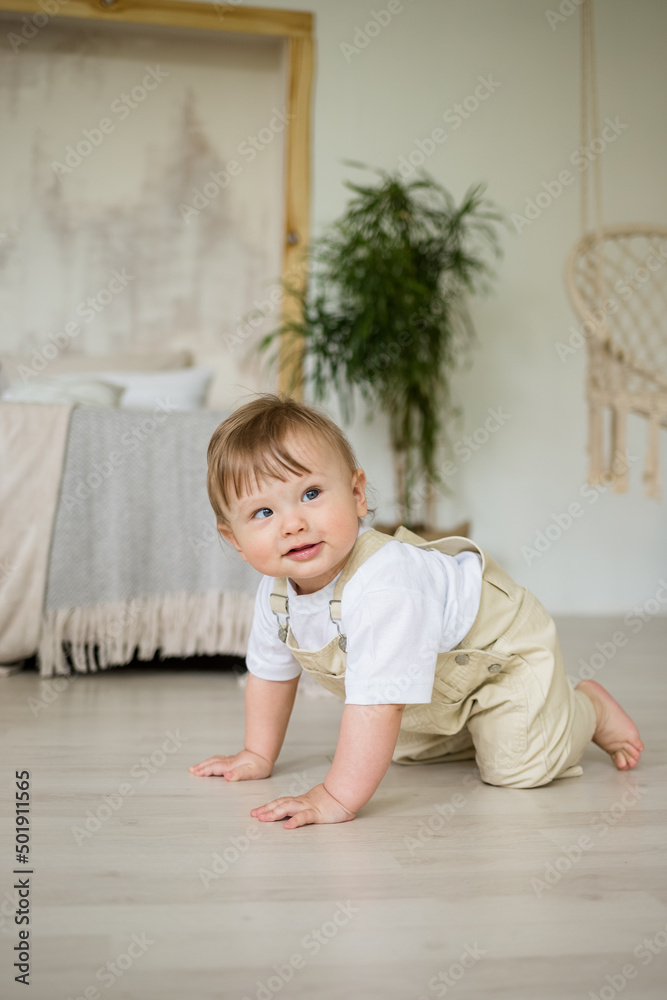 Caucasian baby boy with a squint in a beige jumpsuit crawls on the floor in the bedroom. Child development Caucasian baby boy with a squint in a beige jumpsuit crawls on the floor in the bedroom. Child development