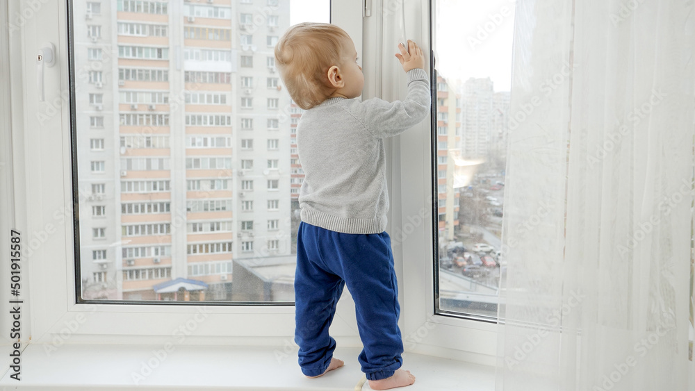 Little baby boy standing on windowsill and pulling window handle. Baby in danger. CHild safety. Little baby boy standing on windowsill and pulling window handle. Baby in danger. CHild safety.