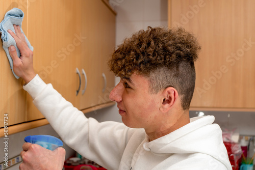 Caucasian young man with curly hair cleaning the kitchen furniture without much enthusiasm, he lives alone and has to do housework photo