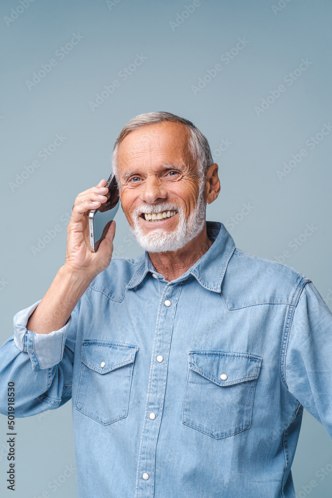 Senior man with grey beard talks to grandchildren on smartphone smiling. Cheerful elderly grandfather enjoys communication on blue background closeup Senior man with grey beard talks to grandchildren on smartphone smiling. Cheerful elderly grandfather enjoys communication on blue background closeup