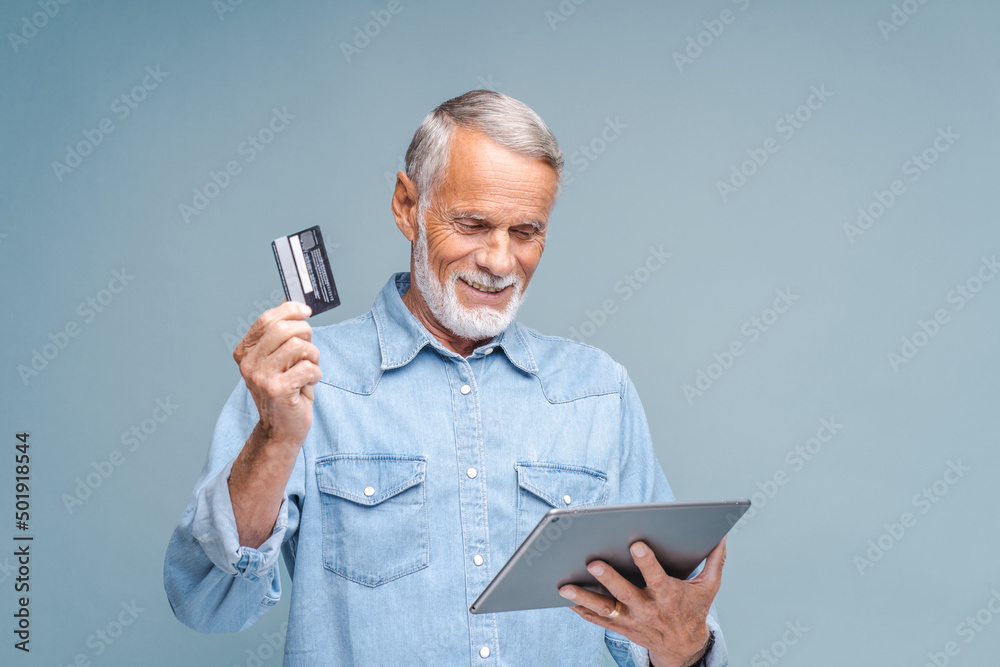 Elderly man makes online purchases on tablet holding credit card with hand. Bearded person in denim shirt stands on grey background looking at gadget Elderly man makes online purchases on tablet holding credit card with hand. Bearded person in denim shirt stands on grey background looking at gadget