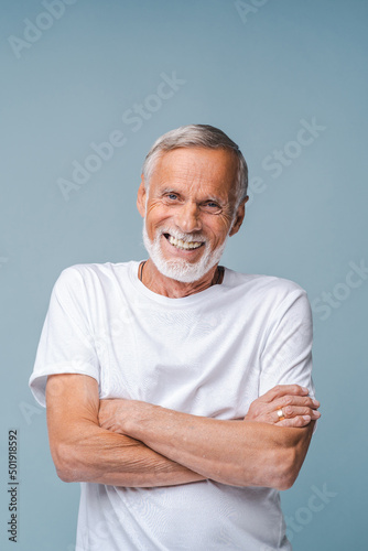 Happy senior man in white t-shirt stands crossing arms. Portrait of bearded elderly pensioner smiling widely on light blue background close view