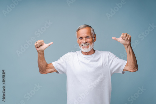 Happy senior man in white t-shirt shows thumbs-up gesture with hands smiling. Portrait of positive bearded grandfather standing on blue background photo