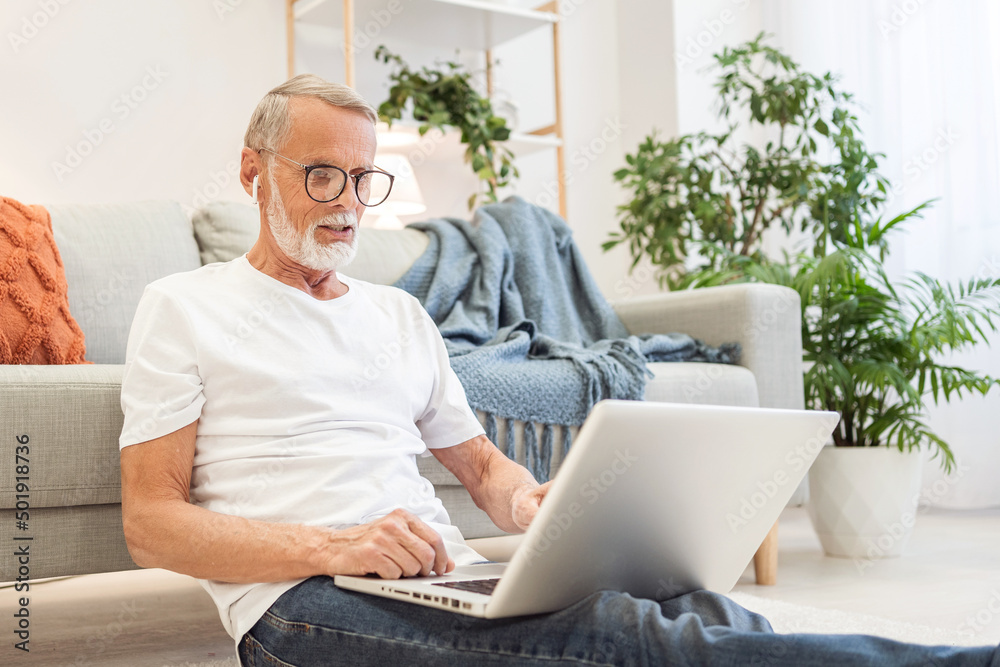 Bearded employee works online in laptop sitting on floor leaning on sofa. Focused senior man in glasses writes report after finishing task at home Bearded employee works online in laptop sitting on floor leaning on sofa. Focused senior man in glasses writes report after finishing task at home