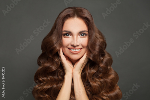 Cheerful young woman with clear skin and curly hair smiling on gray background. Haircare and skincare concept