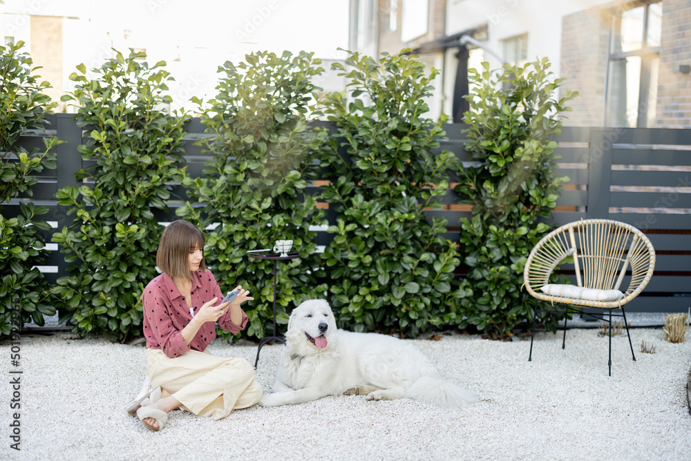 Young woman sitting relaxed with her huge white adorable dog on green hedge background, spending time together at backyard. Friendship with pets and happy leisure time outdoors concept Young woman sitting relaxed with her huge white adorable dog on green hedge background, spending time together at backyard. Friendship with pets and happy leisure time outdoors concept