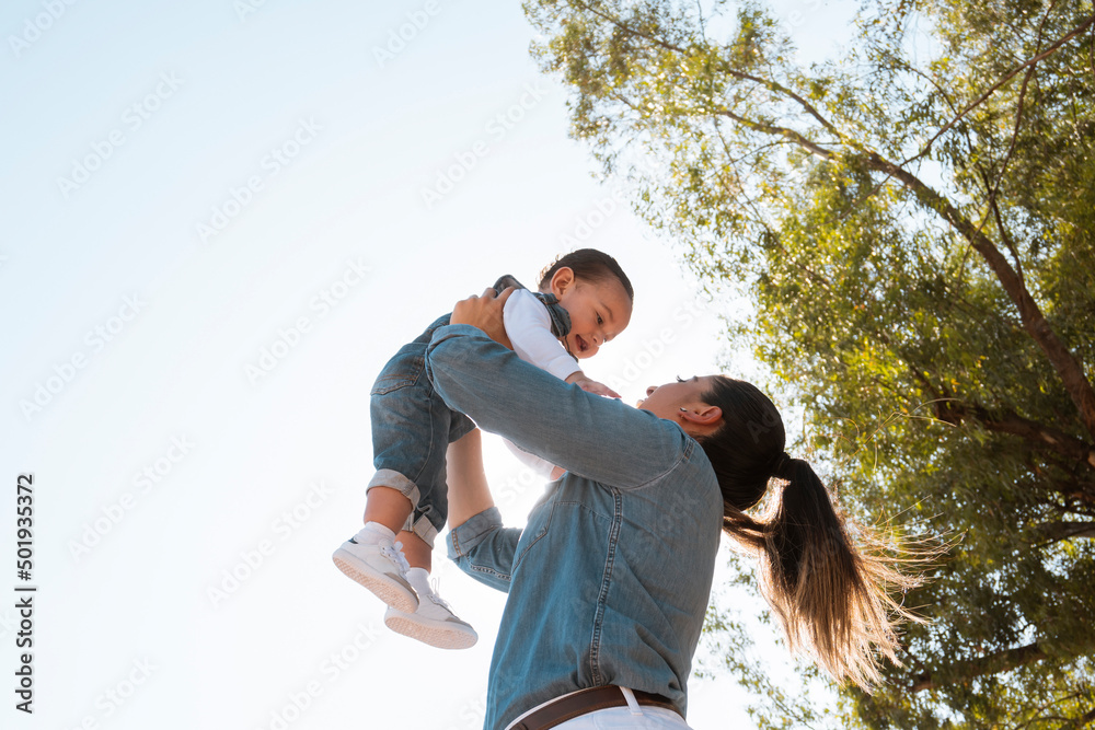 © alejandro - young mother lifts her son in her arms in the park at sunset - low angle