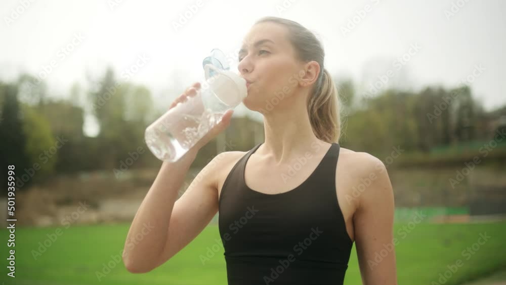 Attractive blond young woman drinking from sport bottle during a workout pause in stadium. Fitness woman drinking water from bottle after working out. . High quality 4k footage Attractive blond young woman drinking from sport bottle during a workout pause in stadium. Fitness woman drinking water from bottle after working out. . High quality 4k footage