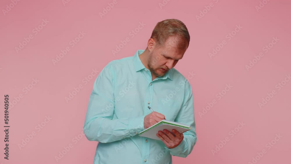 Thoughtful journalist man making notes, writing down thoughts with pen into notepad notebook diary, to do list, good idea. Young adult blonde guy boy isolated alone on pink studio wall background