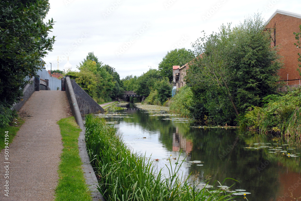 Fototapeta premium Reflections in Calm Waters of Canal with Raised Towpath and Old Mill Building