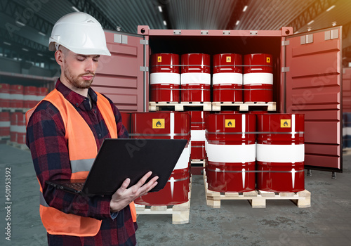 Logistician in warehouse of oil products. Barrels of fuel next to man. Male logistician standing working on laptop. Sea container is filled with barrels of oil. Preparation of oil for export.