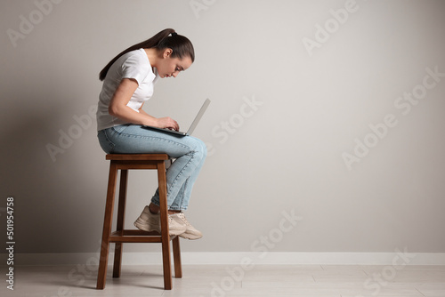 Young woman with poor posture using laptop while sitting on stool near grey wall, space for text