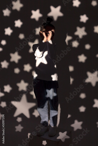 A monochrome soft portrait of a little girl in jeans and a white sweatshirt standing against the wall in a light in a dark room. The girl is smiling on an isolated background. Selective focus