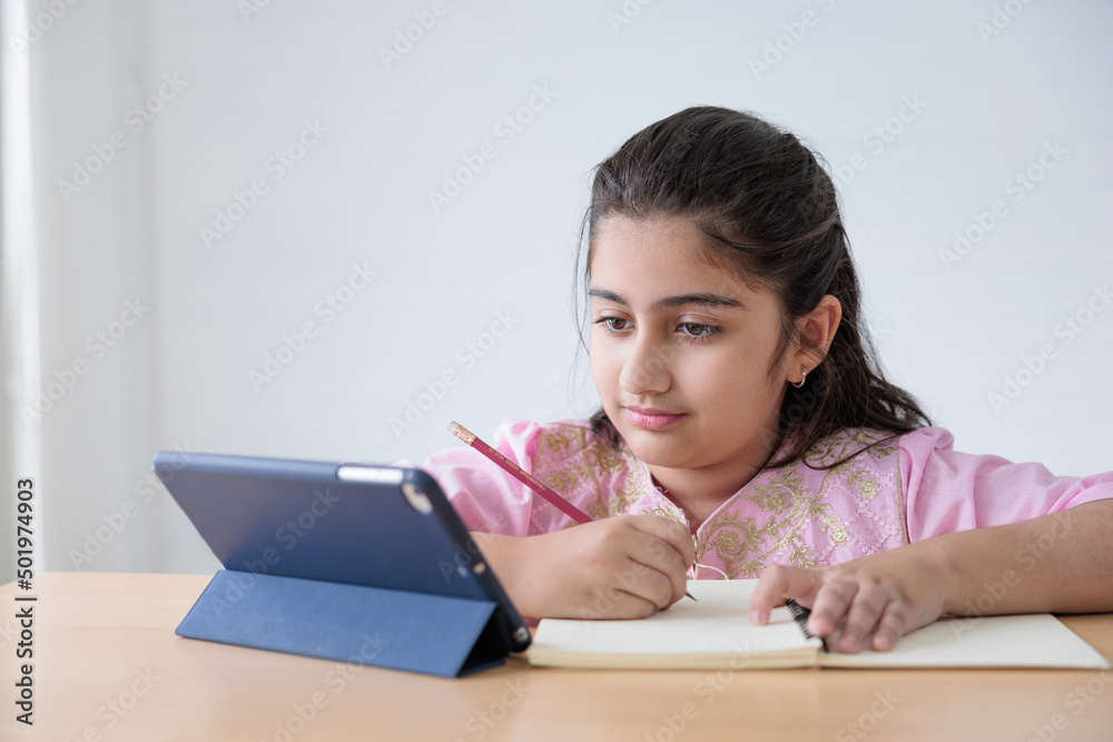 cute Indian girl writing on notebook and looking tablet for online learning class, education concept cute Indian girl writing on notebook and looking tablet for online learning class, education concept