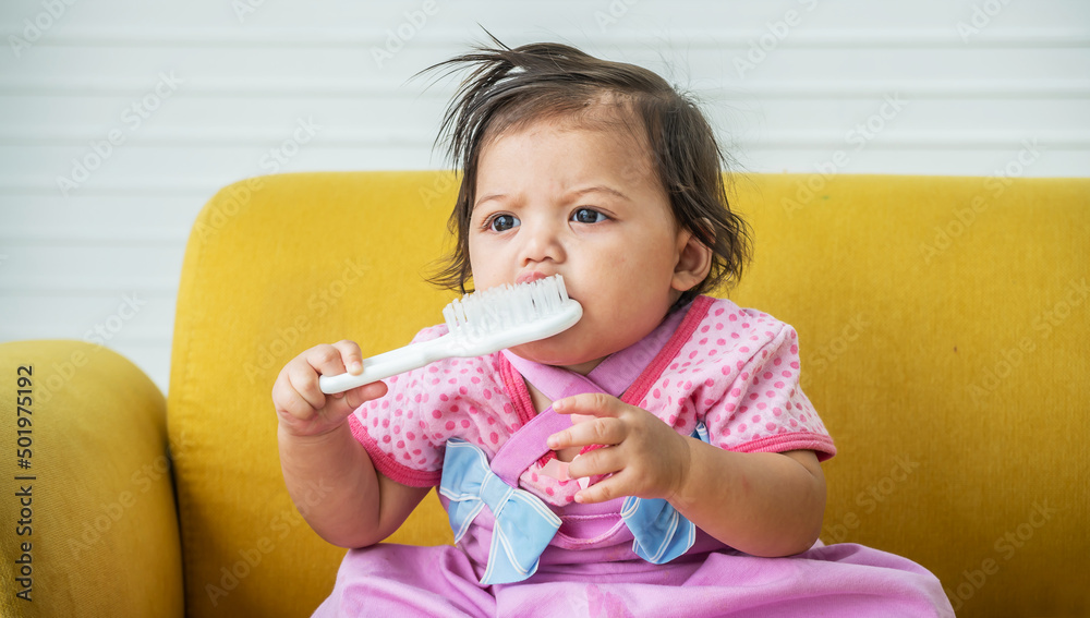 Baby girl is holding and playing a baby hairbrush and smiling. The child sitting biting the hair comb on a sofa. Baby girl is holding and playing a baby hairbrush and smiling. The child sitting biting the hair comb on a sofa.