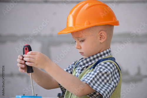 Kid boy twists bolt with screwdriver. Child repairman with repair tool. Child in helmet and boilersuit on construction site. Kids builder and repair.