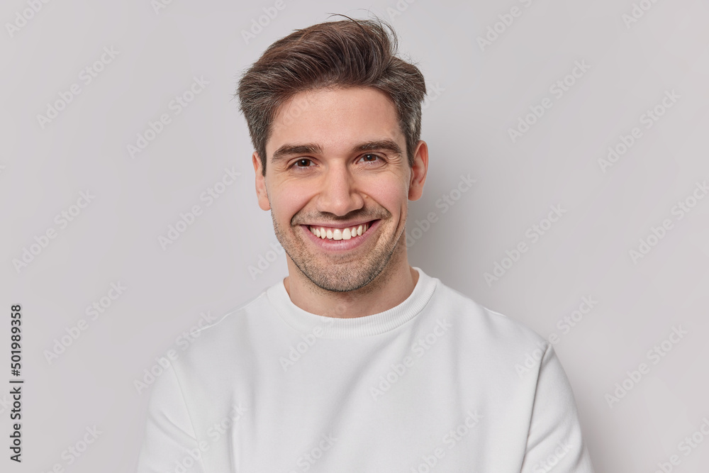 Portrait handsome man with dark hairstyle bristle and toothy smile dressed in white sweatshirt feels very glad poses indoor. Pleased European guy being in good mood smiles positively. Emotions concept Portrait handsome man with dark hairstyle bristle and toothy smile dressed in white sweatshirt feels very glad poses indoor. Pleased European guy being in good mood smiles positively. Emotions concept