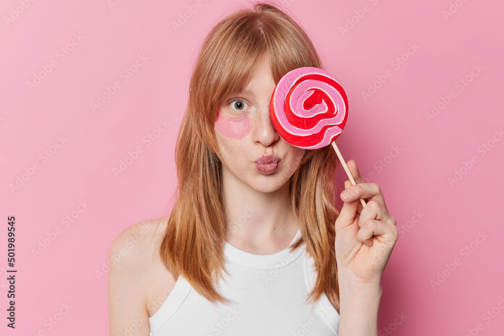 Horizontal shot of young girl with natural ginger haair freckled skin covers caramel candy over eye applies hydrogel patches for reducing puffiness isolated over pink background. Beauty concept