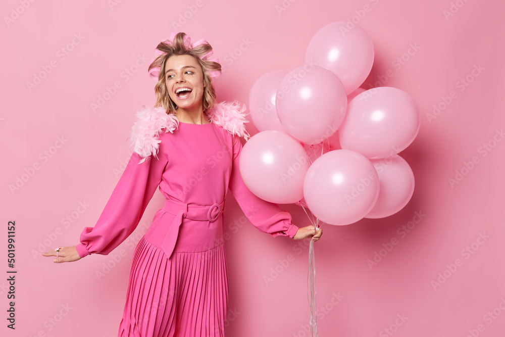 Happy woman wears festive pink dress looks gladfully away applies hair rollers for making hairstyle holds bunch of inflated balloons poses indoor enjoys festive occasion. Celebration concept
