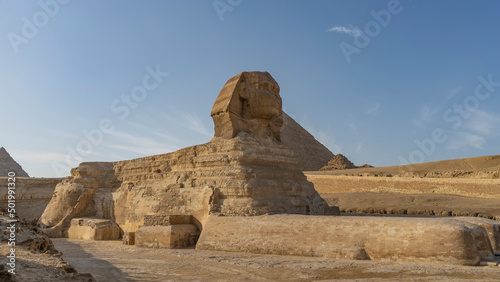Mysterious sculpture of the Great Sphinx. A statue of a mythical creature lying on a plateau. Full-length photo. Paws  tail  and head are visible. Background - blue sky  pyramids. Egypt. Giza
