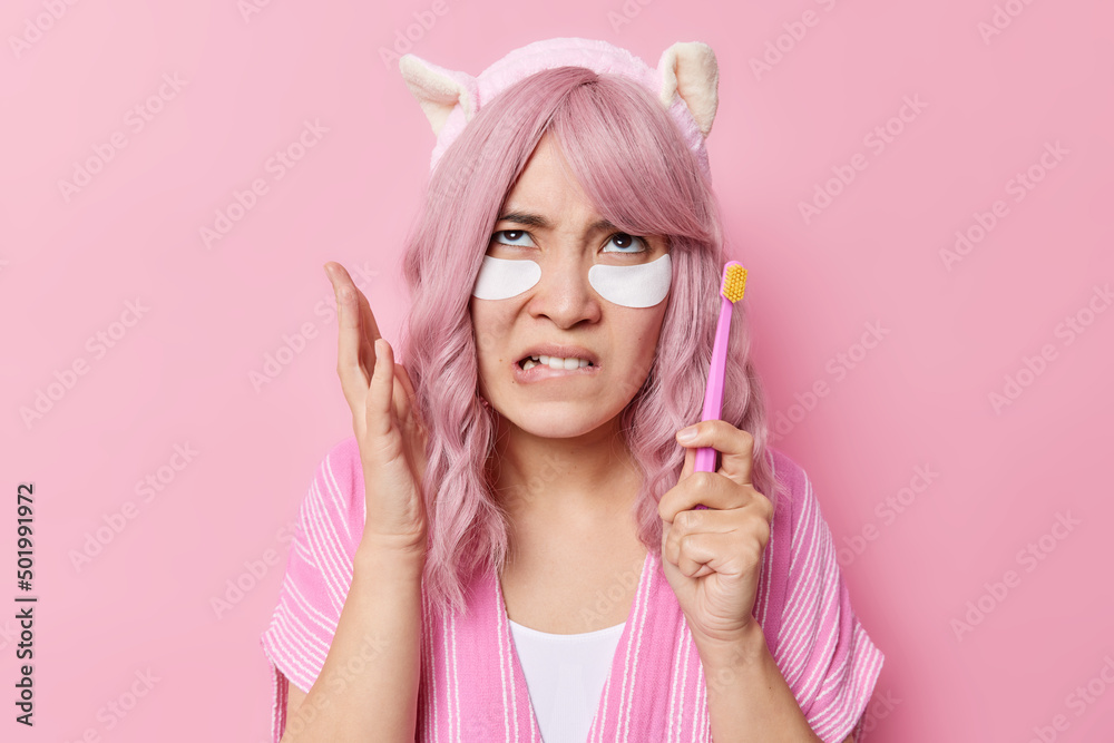 Photo of annoyed displeased Asian woman bites lips focused above holds toothbrush applies beauty patches for skin treatment wears headband isolated over pink background. Daily hygiene routines Photo of annoyed displeased Asian woman bites lips focused above holds toothbrush applies beauty patches for skin treatment wears headband isolated over pink background. Daily hygiene routines