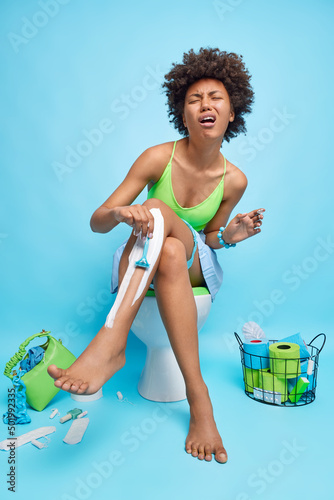 Displeased curly haired crying woman shaves legs undergoes hygiene procedures sits on toilt bowl wears t shirt and skirt isolated over blue background. Women privacy and daily routines concept