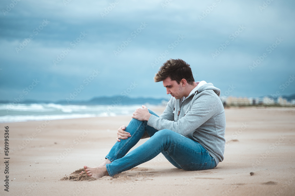 jogging male suffer from a cramp while walking on beach, sitting and massaging the painful muscle.