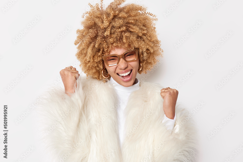 Happy blonde curly haired young woman shakes fists celebrates success makes yes gesture accepts great offer wears spectacles and white fur coat poses indoor achieves goal. Yeah we are champions Happy blonde curly haired young woman shakes fists celebrates success makes yes gesture accepts great offer wears spectacles and white fur coat poses indoor achieves goal. Yeah we are champions