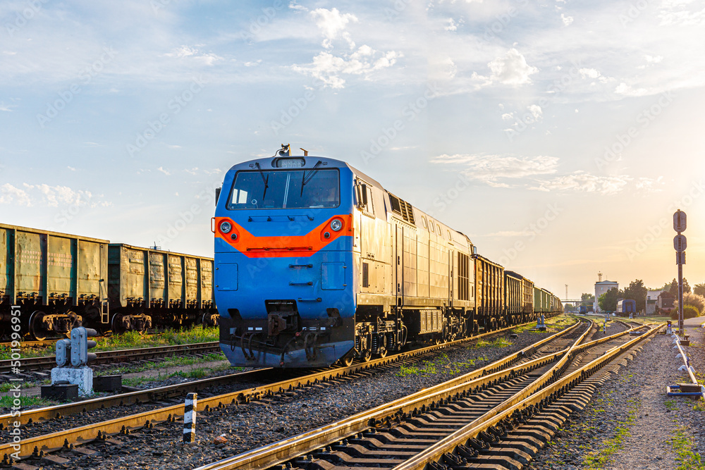 Obraz premium A powerful locomotive with a freight train departs from the marshalling railway station. Sunset lighting.