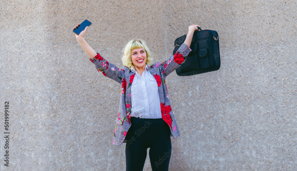 El éxito de la mujer en el mundo laboral,  celebración de los objetivos y metas , mujer feliz con los brazos levantados sujetando un maletín y un teléfono móvil  - obrazy, fototapety, plakaty 