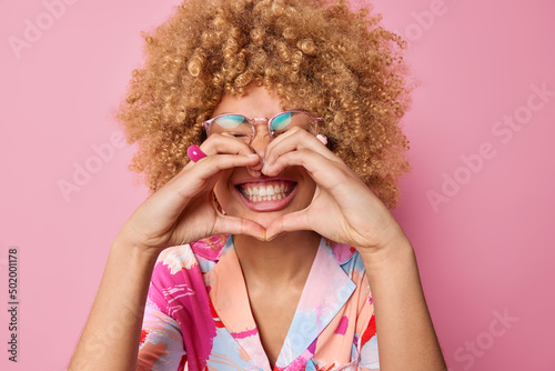 Happy curly haired young woman makes heart gesture over mouth smiles toothily wears transparent spectacles and colorful shirt says I love you isolated over pink background. Body language concept photo