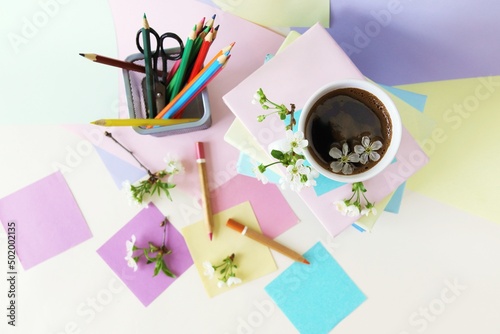 A stack of books, a cup of coffee, pencils, notepads and twigs of cherry blossoms on the table, against a background of colored paper in pastel colors, the concept of learning, education