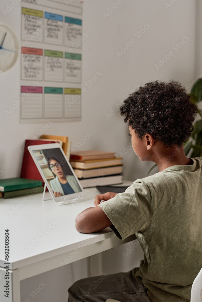 Concentrated Black boy with curly hair sitting at table and using tablet while listening to tutor online Concentrated Black boy with curly hair sitting at table and using tablet while listening to tutor online