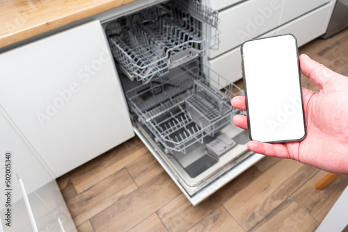 Man holding phone, controlling dishwasher remotely. Smart kitchen appliances concept