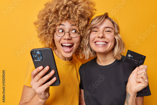 Easy payments. Happy young women make internet shopping use mobile phone and banking card purchase things online smile broadly dressed in casual t shirt isolated over vivid yellow background