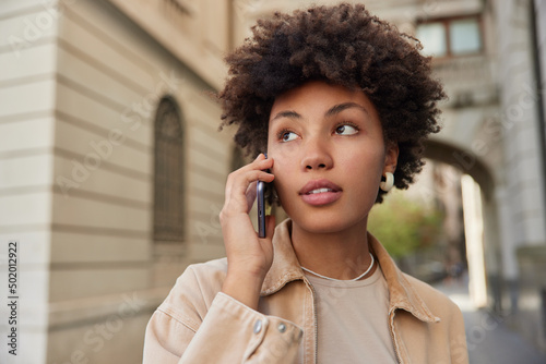 Curly haired beautiful woman has makes cellphone calling strolls outdoors in city wears beige jacket looks away with pensive expression discusses latest news. People lifestyle and leisure concept