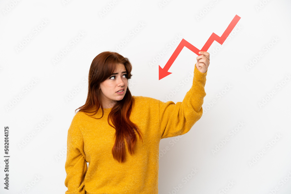 Young redhead woman isolated on white background holding a downward arrow and with sad expression Young redhead woman isolated on white background holding a downward arrow and with sad expression