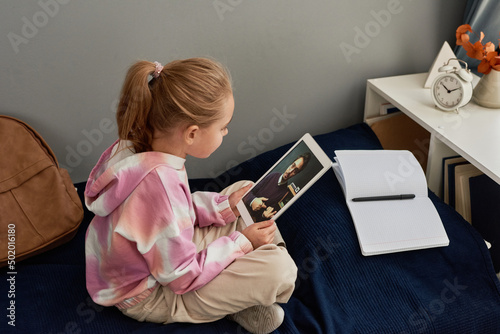 Concentrated girl with ponytale sitting on bed with open workbook and using tablet for communication with tutor photo