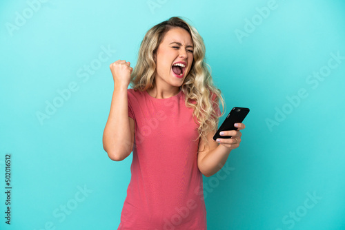 Young Brazilian woman isolated on blue background using mobile phone and doing victory gesture