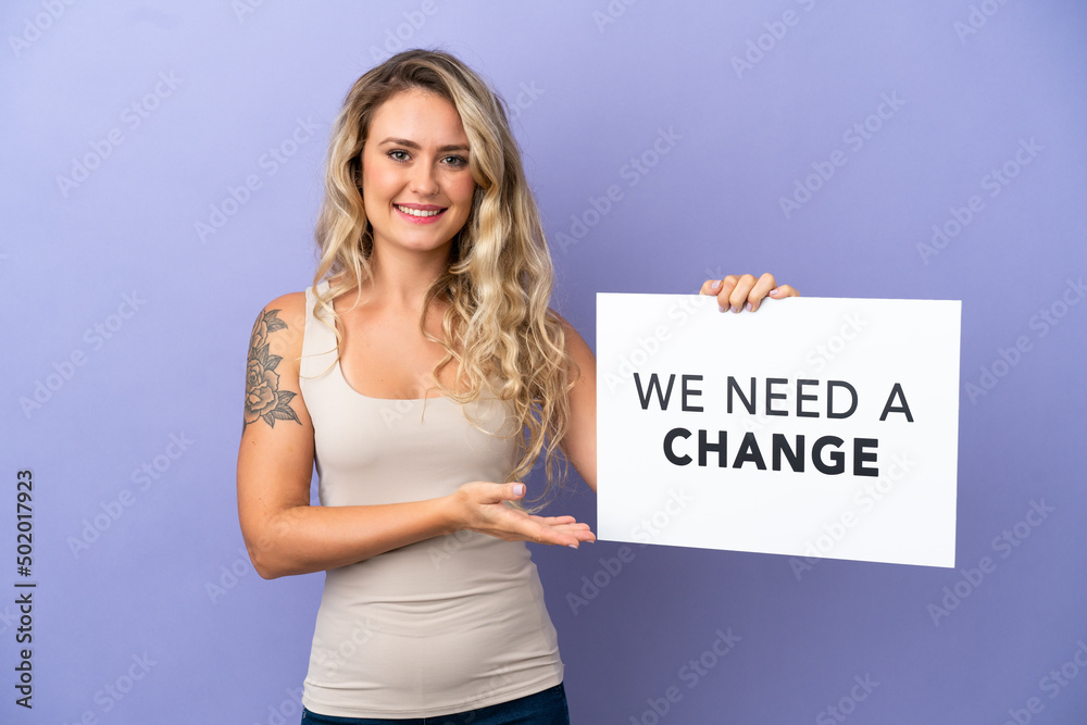 Young Brazilian woman isolated on purple background holding a placard with text We Need a Change and pointing it Young Brazilian woman isolated on purple background holding a placard with text We Need a Change and pointing it