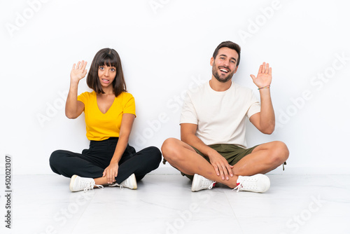 Young couple sitting on the floor isolated on white background saluting with hand with happy expression