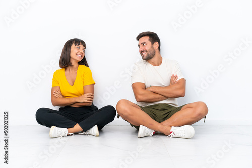 Young couple sitting on the floor isolated on white background looking over the shoulder with a smile