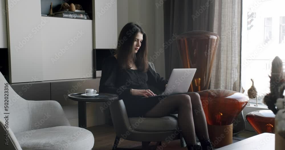 Pretty young Caucasian woman texting on the keyboard of the laptop computer and sitting on the sofa in the cozy living room. Indoors. Home leisure. Indoors relaxing. Writing hobby. Online job at home. Pretty young Caucasian woman texting on the keyboard of the laptop computer and sitting on the sofa in the cozy living room. Indoors. Home leisure. Indoors relaxing. Writing hobby. Online job at home.