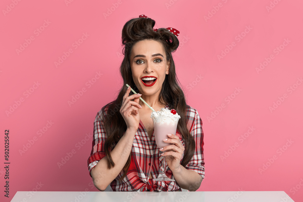 Excited young pinup woman in retro style clothes drinking milk shake from glass, sitting at table over blue background Excited young pinup woman in retro style clothes drinking milk shake from glass, sitting at table over blue background