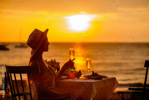 Romantic dinner near sea on sunset. Woman sitting alone, waiting husband on table set for a romantic meal on beach sky and ocean on background. Dinner for a couple in love in luxury outdoor restaurant photo