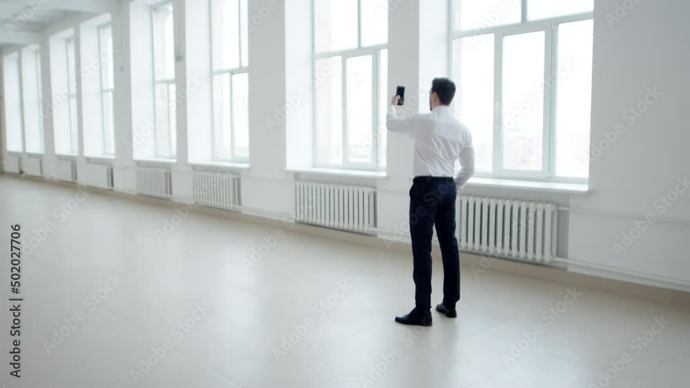 Male realtor stands in middle of an empty office space and shoots it on smartphone, back view. Man stands and takes off an empty room on smartphone Male realtor stands in middle of an empty office space and shoots it on smartphone, back view. Man stands and takes off an empty room on smartphone