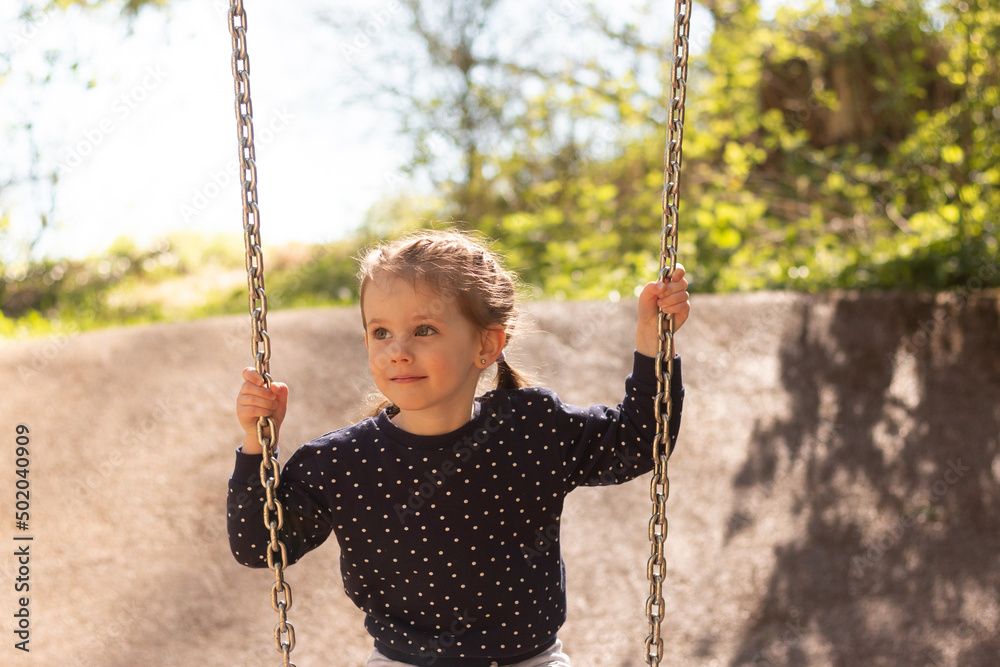 A little beautiful girl with pigtails, smiling, rides on a swing in polka-dot clothes in a park against the background of foliage A little beautiful girl with pigtails, smiling, rides on a swing in polka-dot clothes in a park against the background of foliage