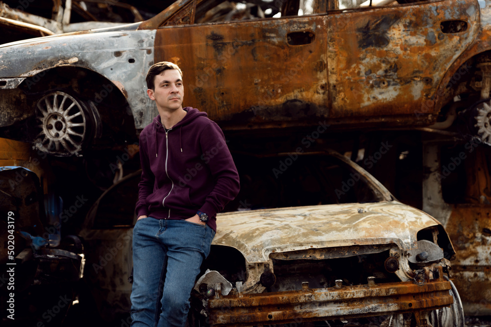 A Ukrainian from Kyiv stands next to broken and burnt cars that were damaged by rocket explosions. war in Ukraine. Russian aggression in Ukraine. a man in front of a burned-out car. A Ukrainian from Kyiv stands next to broken and burnt cars that were damaged by rocket explosions. war in Ukraine. Russian aggression in Ukraine. a man in front of a burned-out car.