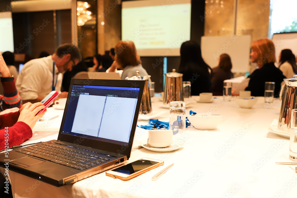 desktop computer, mobile phone, glass, water bottle on a table with blurred meeting participants and screen in background in a conference hall. desktop computer, mobile phone, glass, water bottle on a table with blurred meeting participants and screen in background in a conference hall.